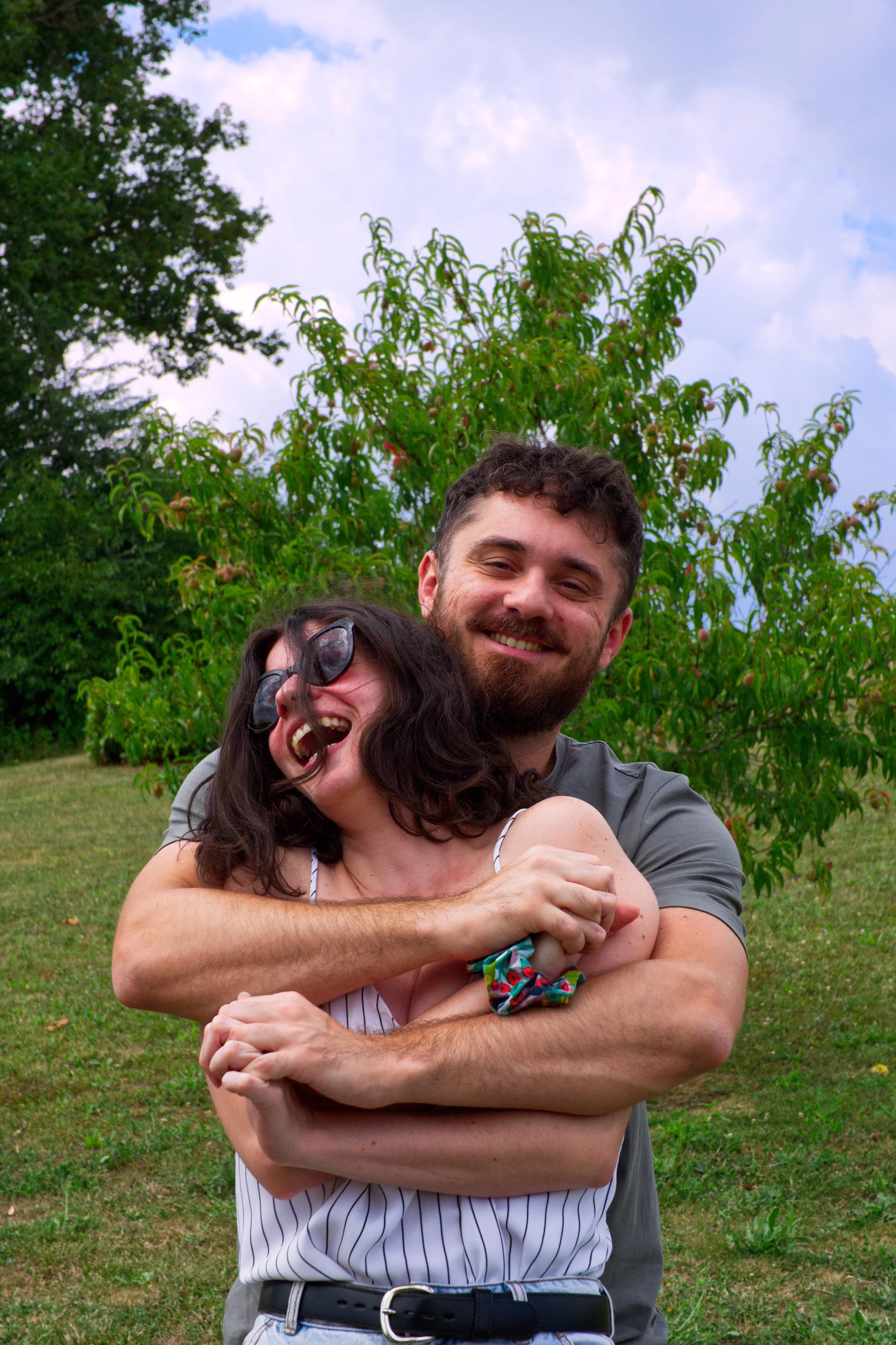Deux personnes souriantes, un homme et une femme, s'enlacent en plein air avec des arbres et un ciel nuageux en arrière-plan. Portraits duo