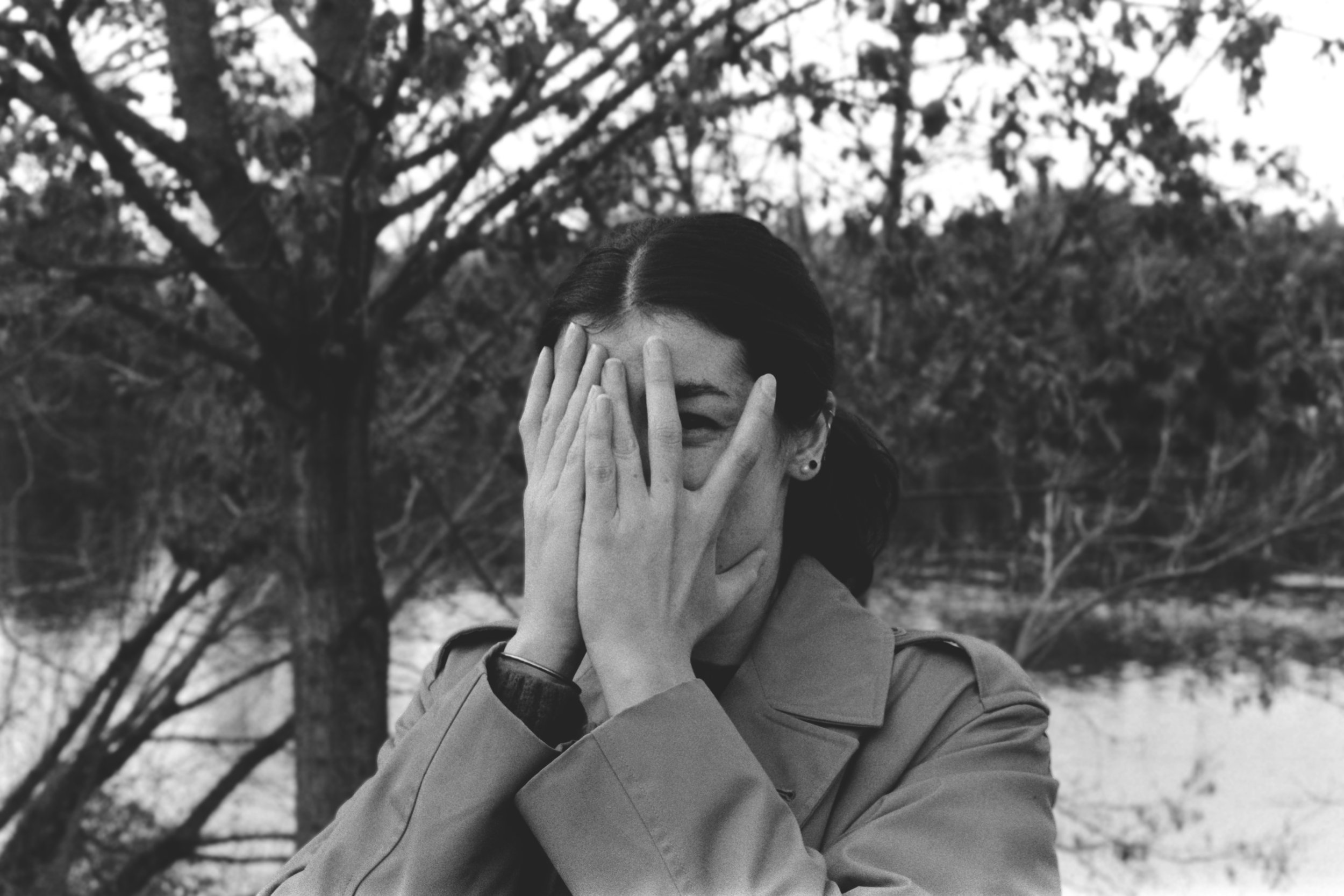 Une femme en manteau, se couvrant partiellement le visage avec sa main, se tient devant un arbre dans un paysage naturel en noir et blanc. Portrait