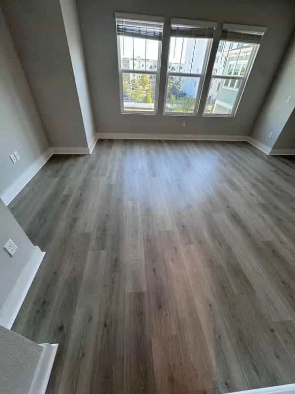 Empty room with hardwood flooring and three large windows with blinds, overlooking neighboring buildings and trees. Own Style Flooring