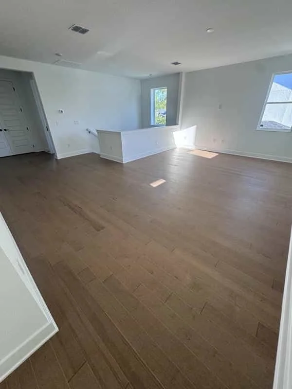 Empty living room with light hardwood floors, white walls, three windows allowing natural light, and a railing on a small step. Own Style Flooring