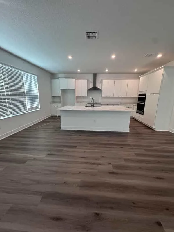Modern kitchen with white cabinets, a large island with a marble countertop, dark wood flooring, and a large window with blinds. Own Style Flooring