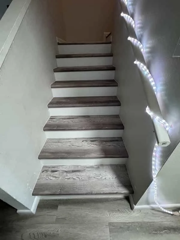 Interior view of a staircase with wooden steps and a light-colored wall on one side, decorated with a string of white lights along the edge. Own Style Flooring