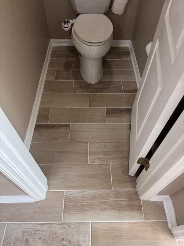 View of a toilet with beige walls, wood-look tile flooring, and white baseboards in a small bathroom, with a white door partially open. Own Style Flooring