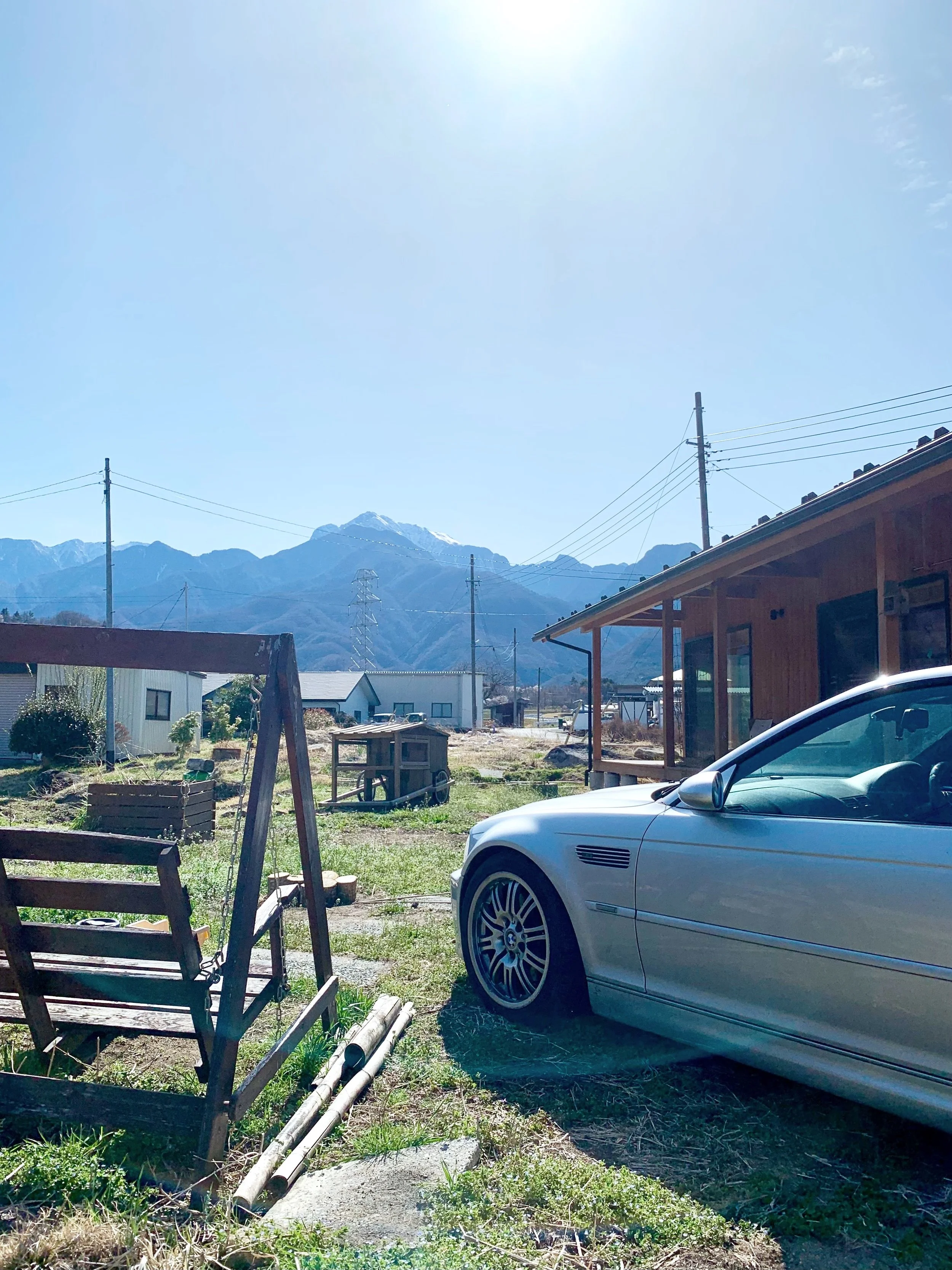庭に駐車された銀色の車、木製のベンチ、遠くに山々と青空が見える風景。