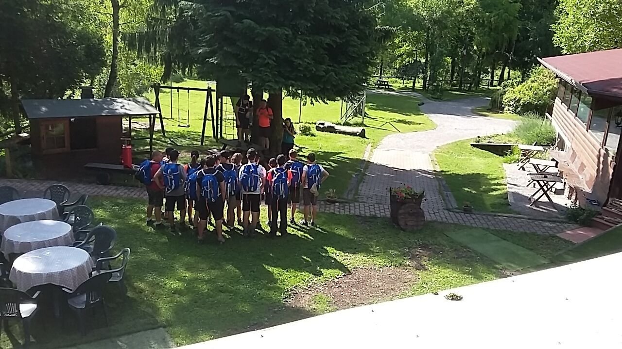Gruppo di ragazzi con zaini blu che ascolta istruzioni sotto un albero in un giardino