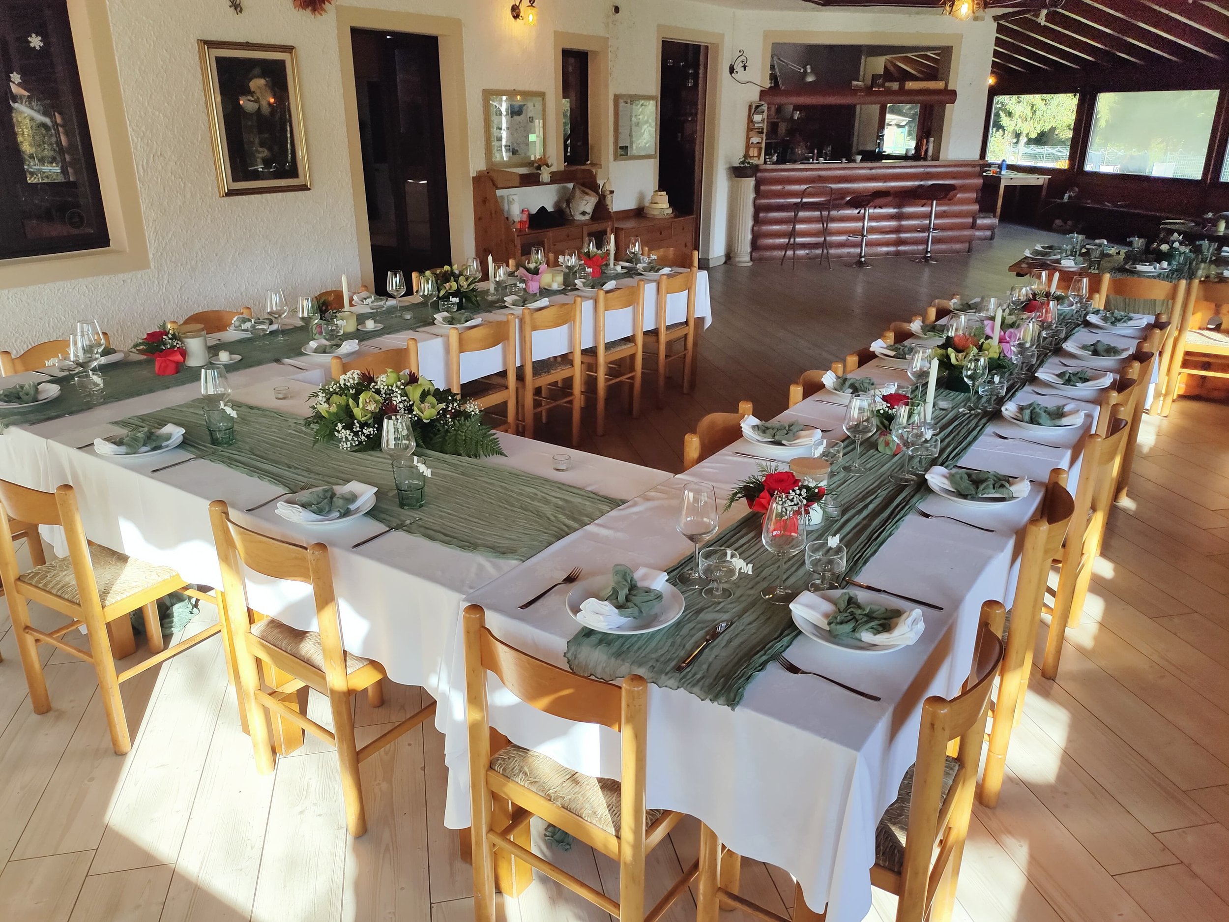 Una sala da pranzo decorata con fiori e candele, con un lungo tavolo apparecchiato con piatti, bicchieri e tovaglioli, pronta per un evento o una cena speciale, con pareti bianche e grandi finestre che lasciano entrare luce naturale.