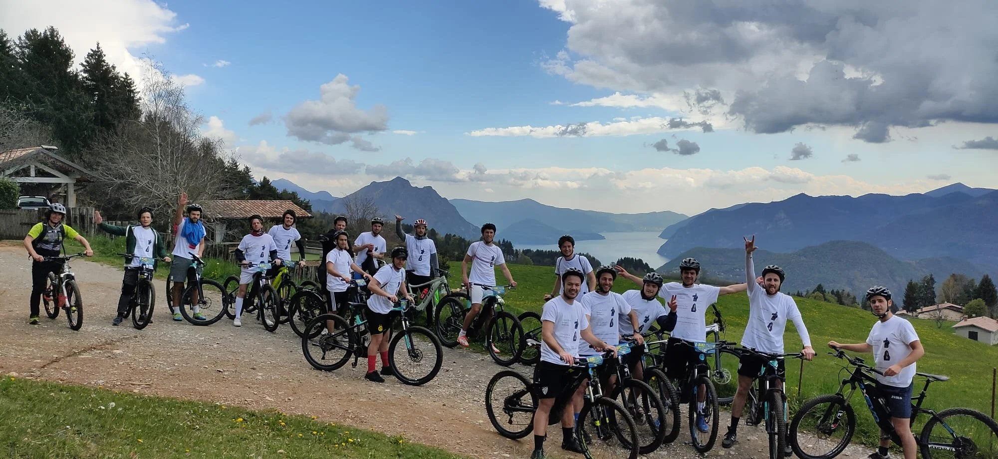 Gruppo di ciclisti con caschi e magliette bianche, alcune persone fanno il gesto della vittoria, con uno sfondo di montagne e lago, in un paesaggio naturale.