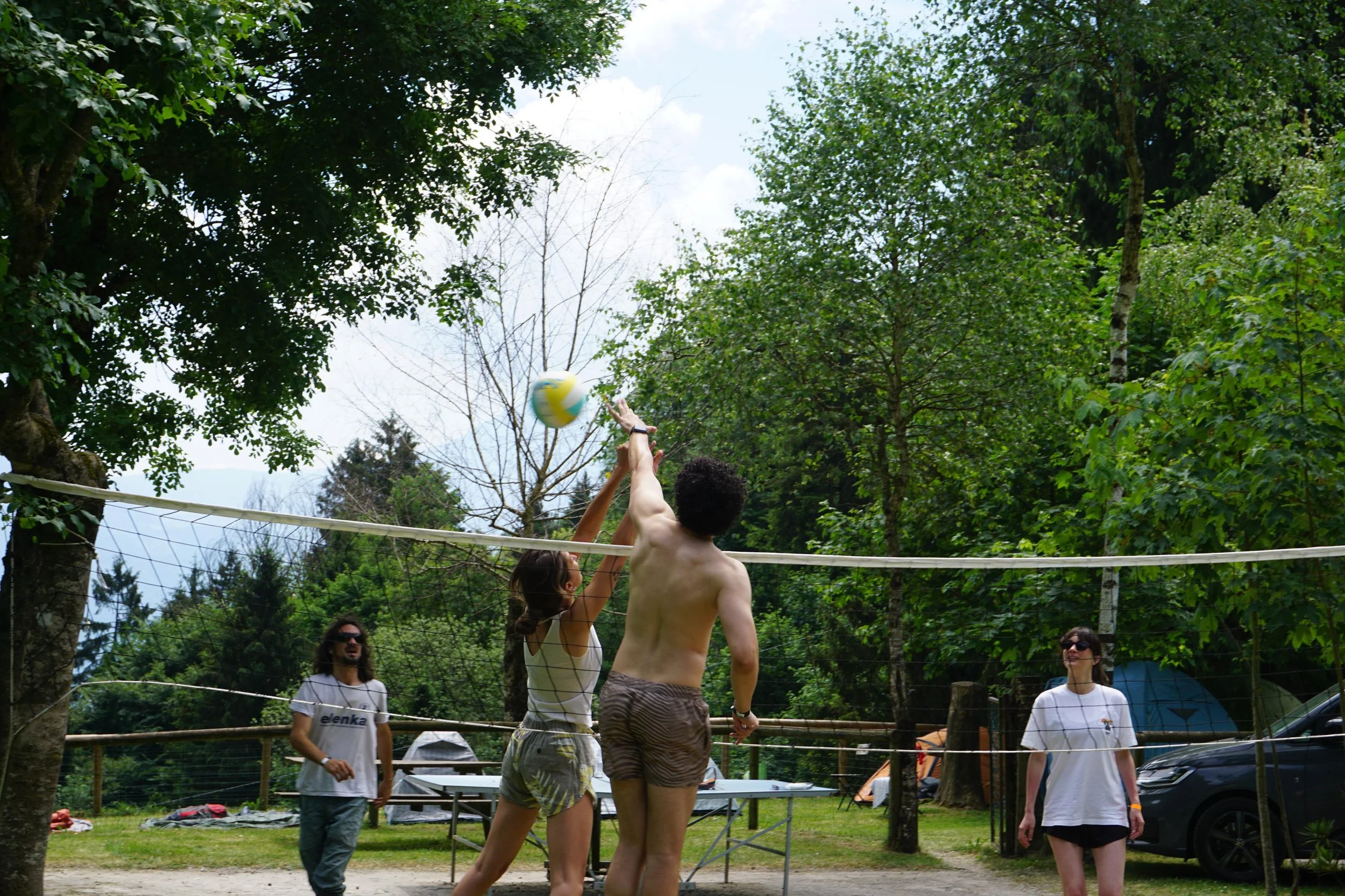 Persone che giocano a pallavolo su un campo all'aperto, circondato da alberi e persone che osservano.