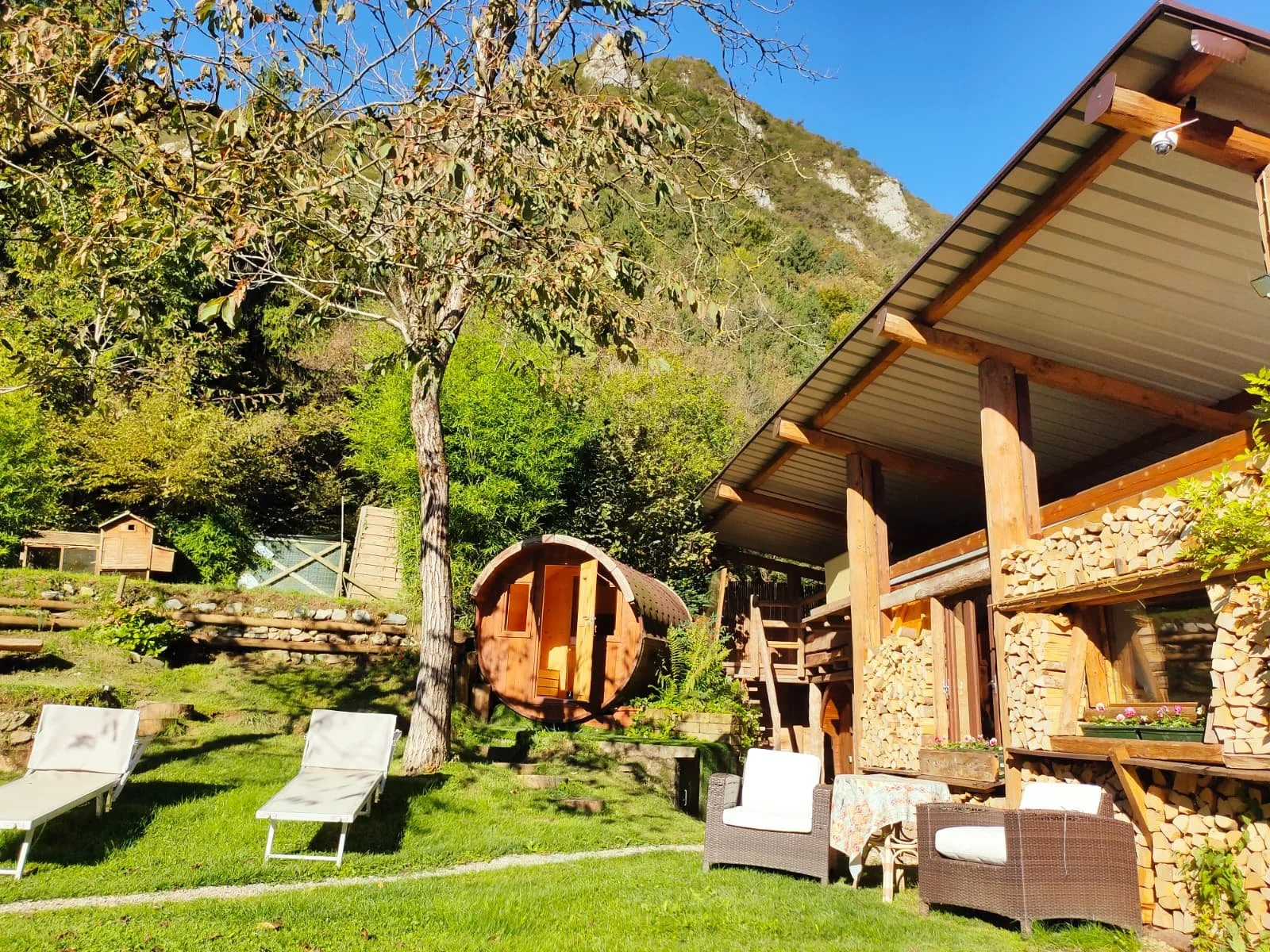 Giardino soleggiato con sedie a sdraio, alberi e casa in legno con pile di legna e un'area relax, alti montagne e cielo blu sullo sfondo.