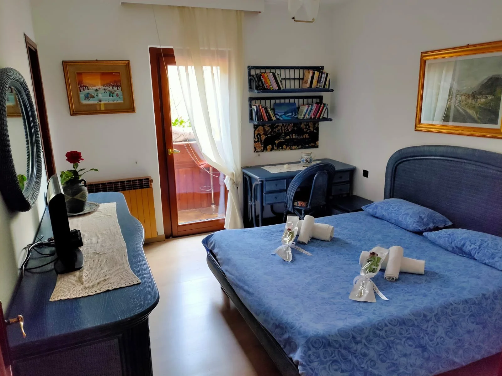 Camera da letto con letto matrimoniale coperto da una coperta blu, decorato con cinque decorazioni in fiore e asciugamani arrotolati. Pareti bianche con quadri alle pareti, una scrivania blu con sedia, una libreria sopra la scrivania, e una porta-fin