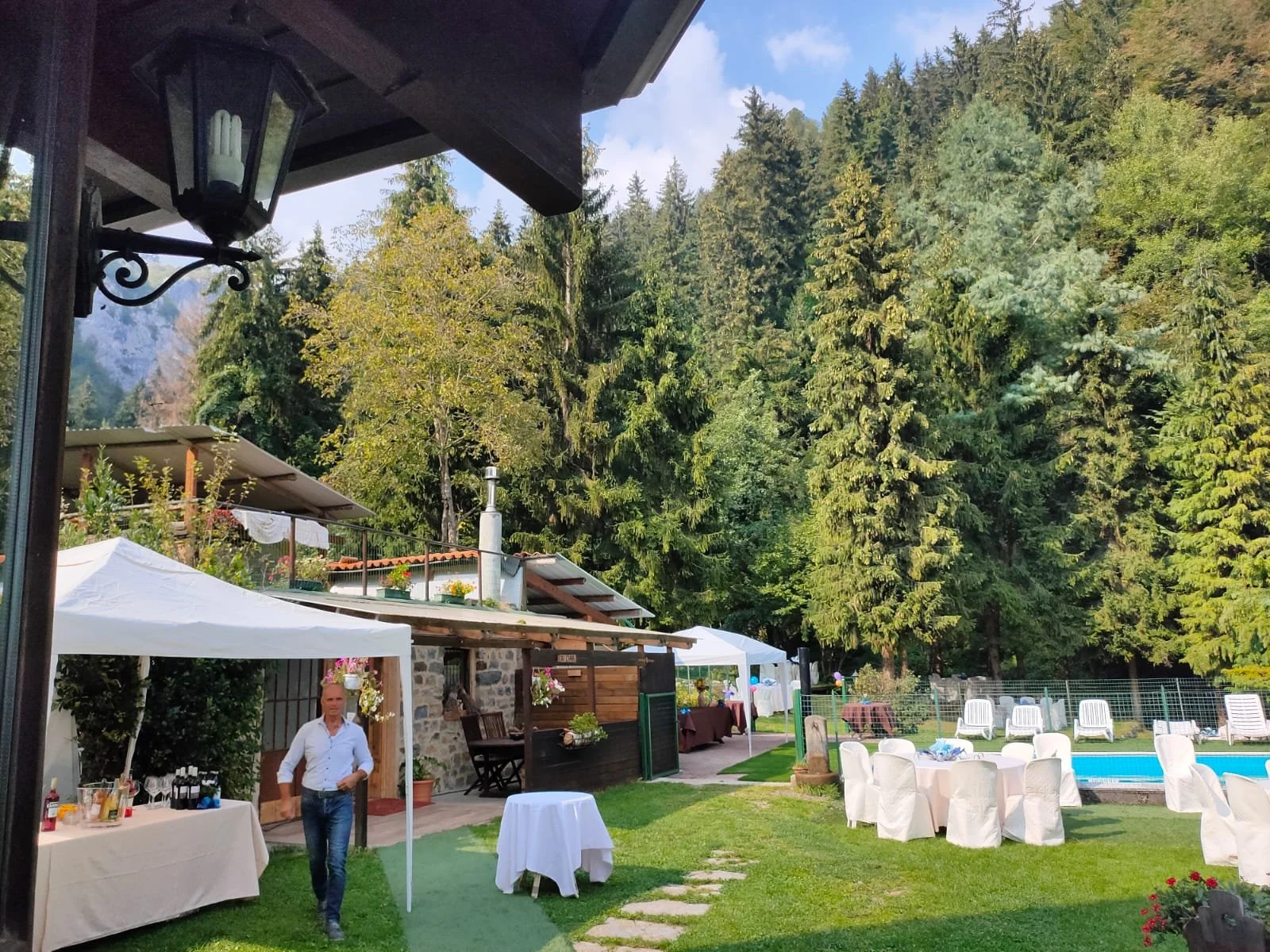 Giardino all'aperto con tavoli e sedie, tende, persone, piscina, alte piante e alberi, ambientazione per evento o festa