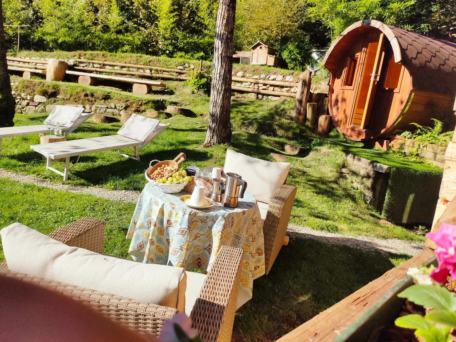 Zona relax all'aperto con sedie, tavolo con bevande, frutta, e ambiente boschivo con casetta di legno e panchine in legno