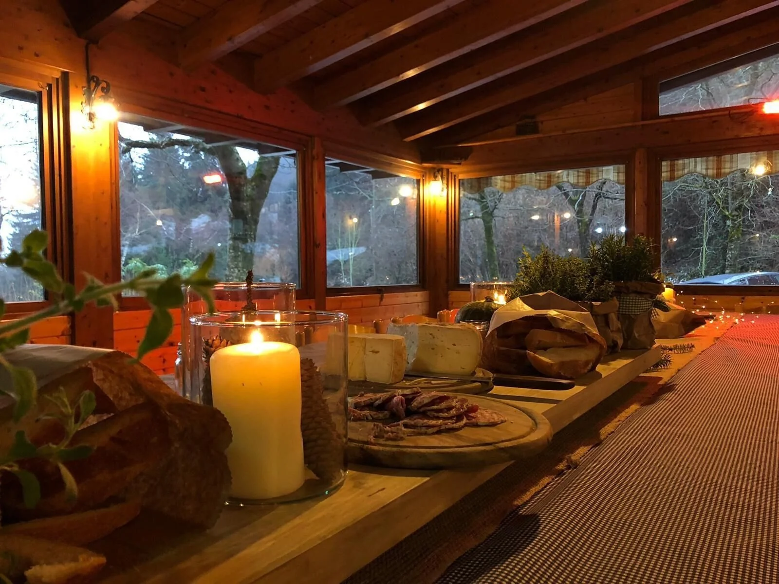 Interno di una stanza in legno con grandi finestroni che mostrano un paesaggio boscoso, decorata con candele accese, una tavola con formaggi, pane, salumi e piante.