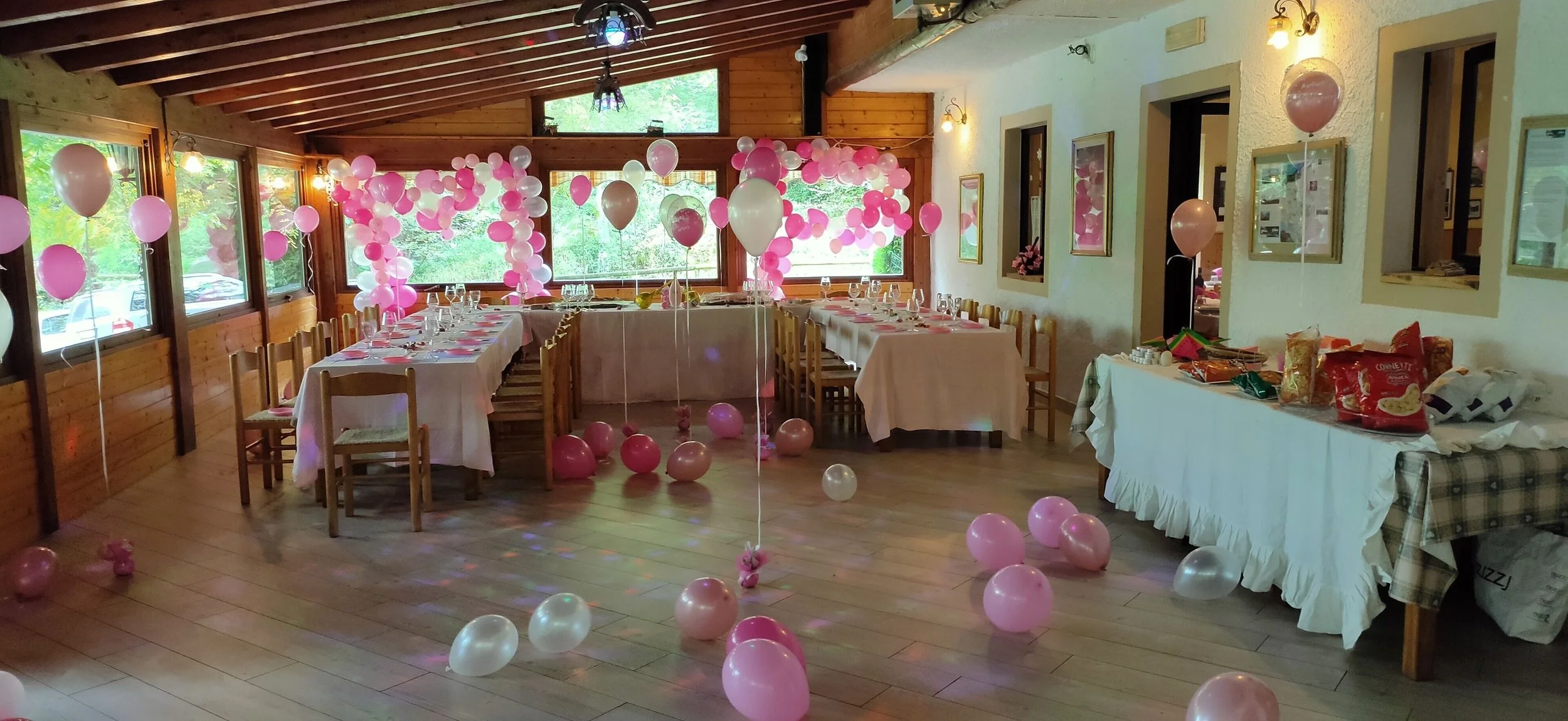 Sala decorata per festa con molte decorazioni di palloncini rosa e bianchi, tavoli apparecchiati per un ricevimento, con finestra che mostra il verde esterno, e alcune scatole e sacchetti sulla destra.