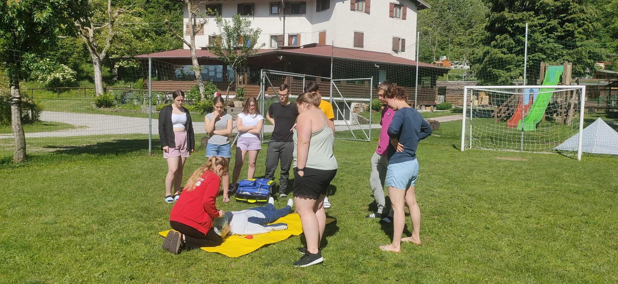 Gruppo di persone mentre partecipano a un corso di pronto soccorso su un prato verde con a fianco un'area gioco per bambini e un'area residenziale sullo sfondo.