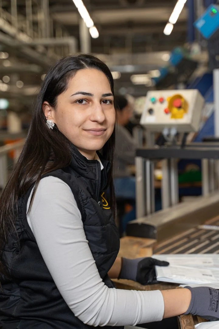 Eine junge Frau mit dunklen Haaren, die bei der Österreichischen Post arbeitet, sitzt an einem Förderband mit Papieren in den Händen, trägt schwarze Handschuhe und einen schwarzen Steppweste.