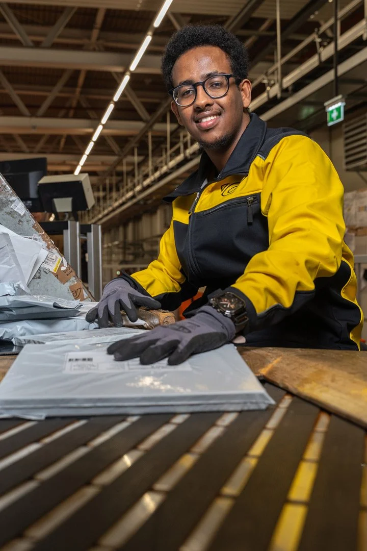 Ein junger Mann arbeitet iim Briefverteilerzentrum der Österreichischen Post. Er trägt eine gelb-schwarze Jacke, schwarze Brille und Handschuhe, und hat Papiere auf dem Fließband.