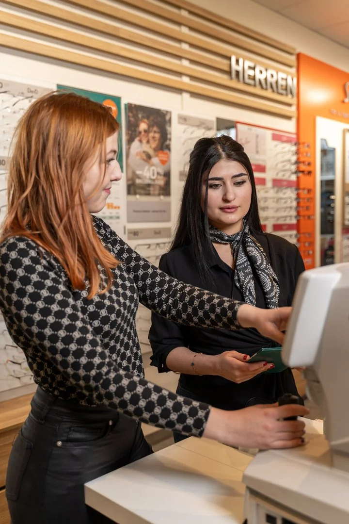 Zwei Frauen am Optikgerät von Pearl. Sie befinden sich in der Filiale, wo Brille hinter ihnen im Regal sind.