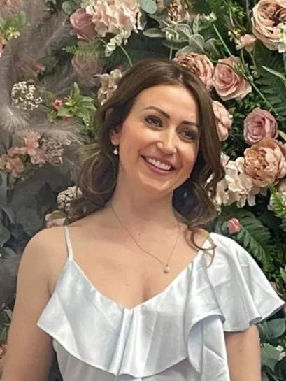 Woman smiling in front of a floral backdrop, wearing a light blue dress with ruffles.
