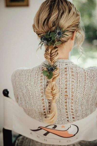 Frau mit Fischgrätenzopf und Blumendekoration im Haar, sitzt auf einem Stuhl; trägt einen gestrickten Pullover.