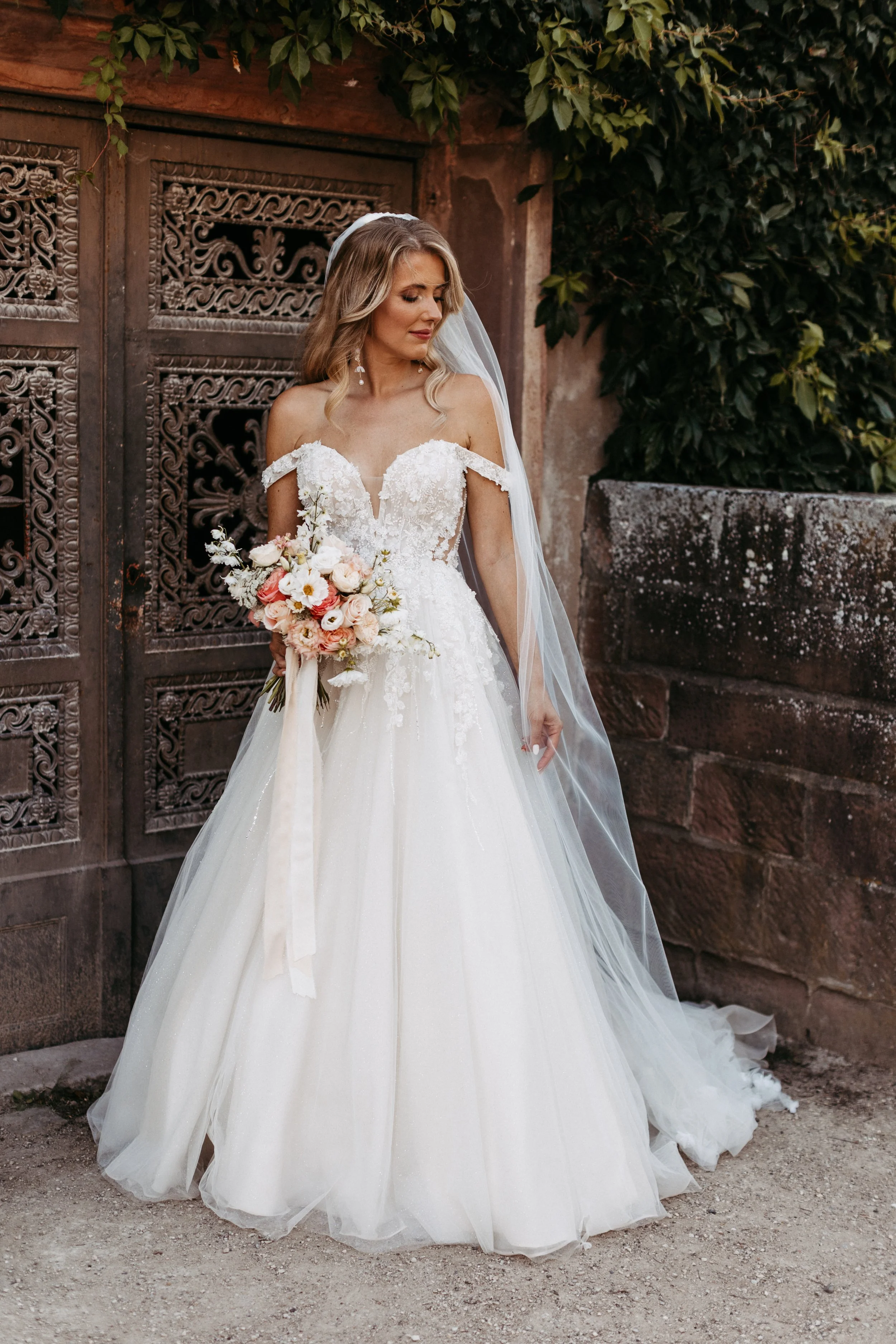 Braut mit Blumenstrauß in der hand vor altem Tor im weißen Kleid und Brautkleid