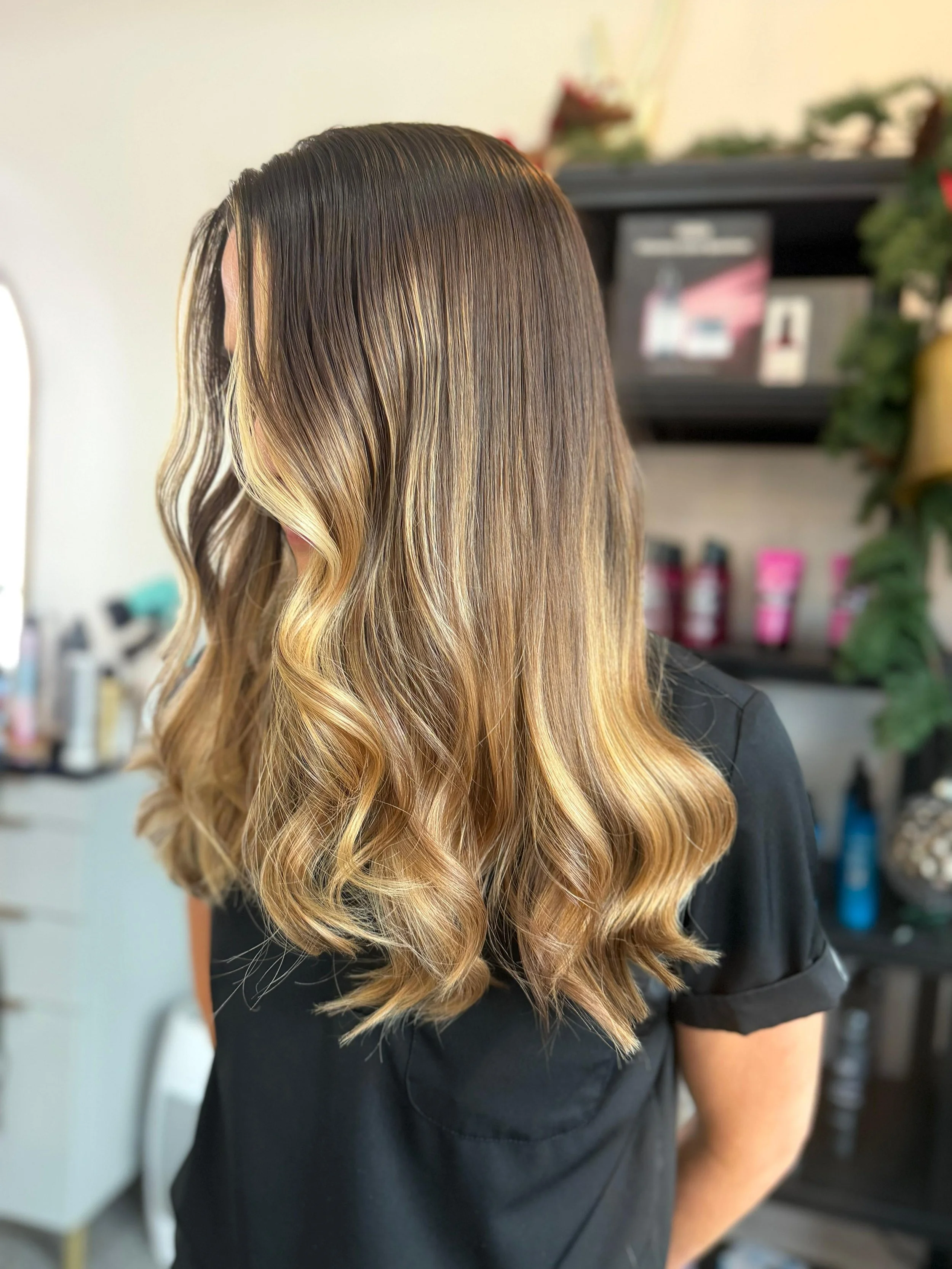 A woman with shoulder-length wavy hair styled with dark roots fading into caramel and honey blonde highlights, standing in a salon.