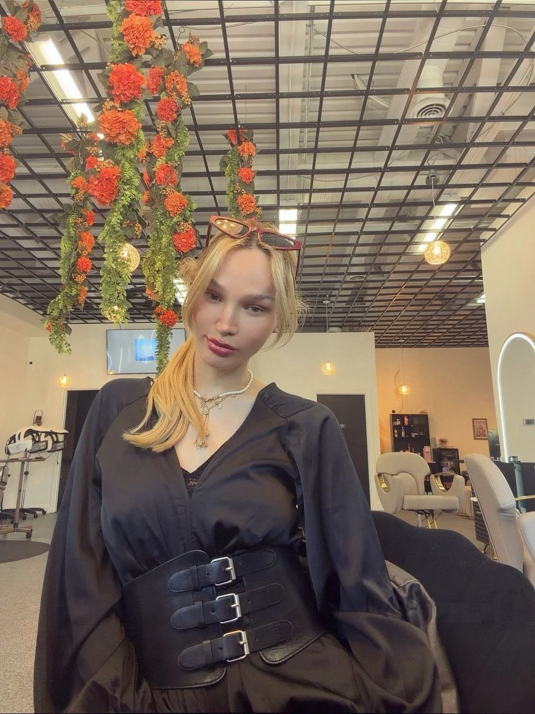A woman with long blonde hair in a white dress standing at a reception desk with a sign that reads "Boho Beauty". She is holding a pair of sunglasses and smiling. The reception area has decorative items, including bottles and a bowl, and is inside a stylish, well-lit space.