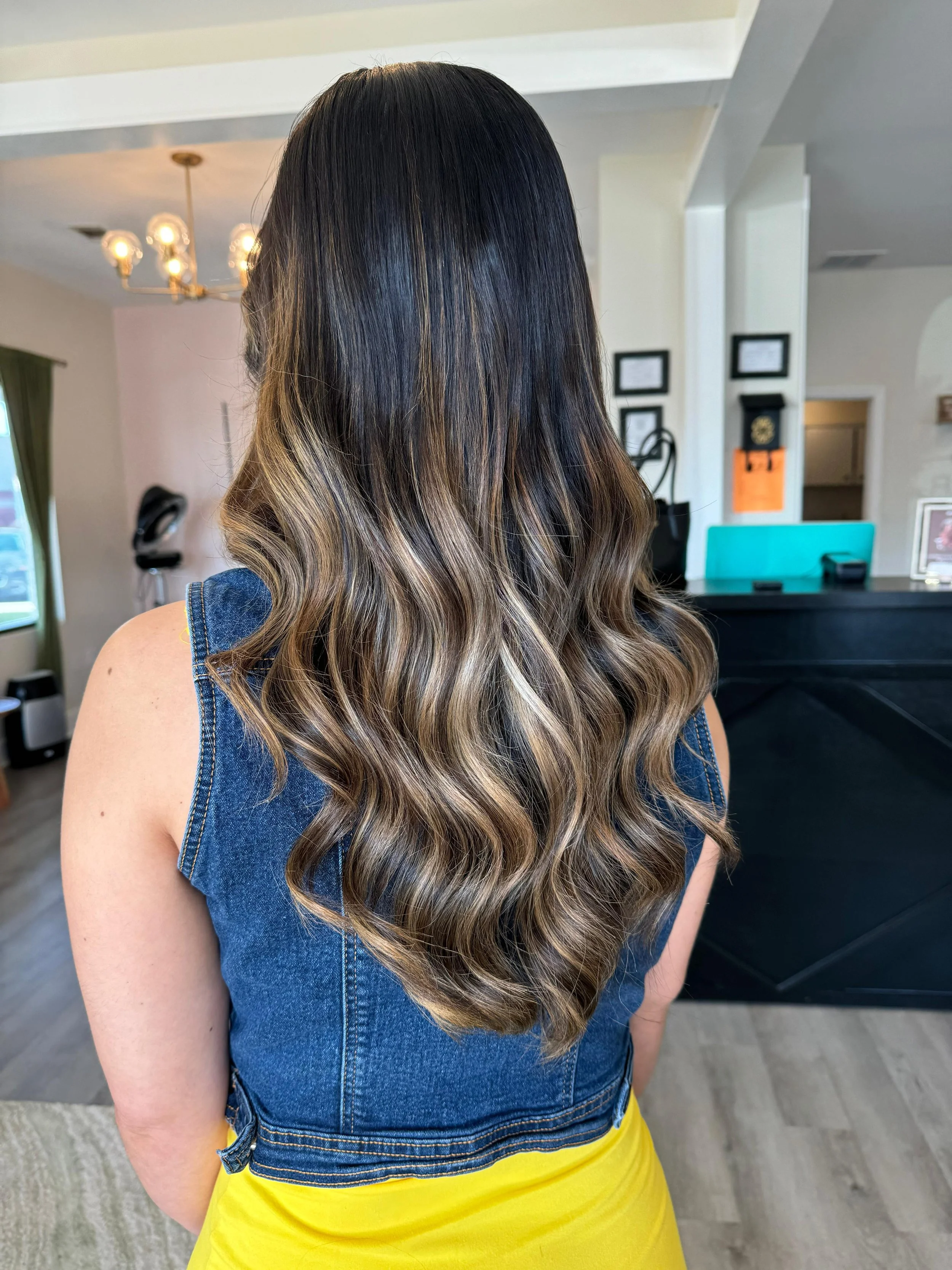 Back view of a woman with long, wavy hair styled in dark brown to blonde balayage, wearing a sleeveless denim vest and a yellow skirt, standing inside a salon or similar setting.