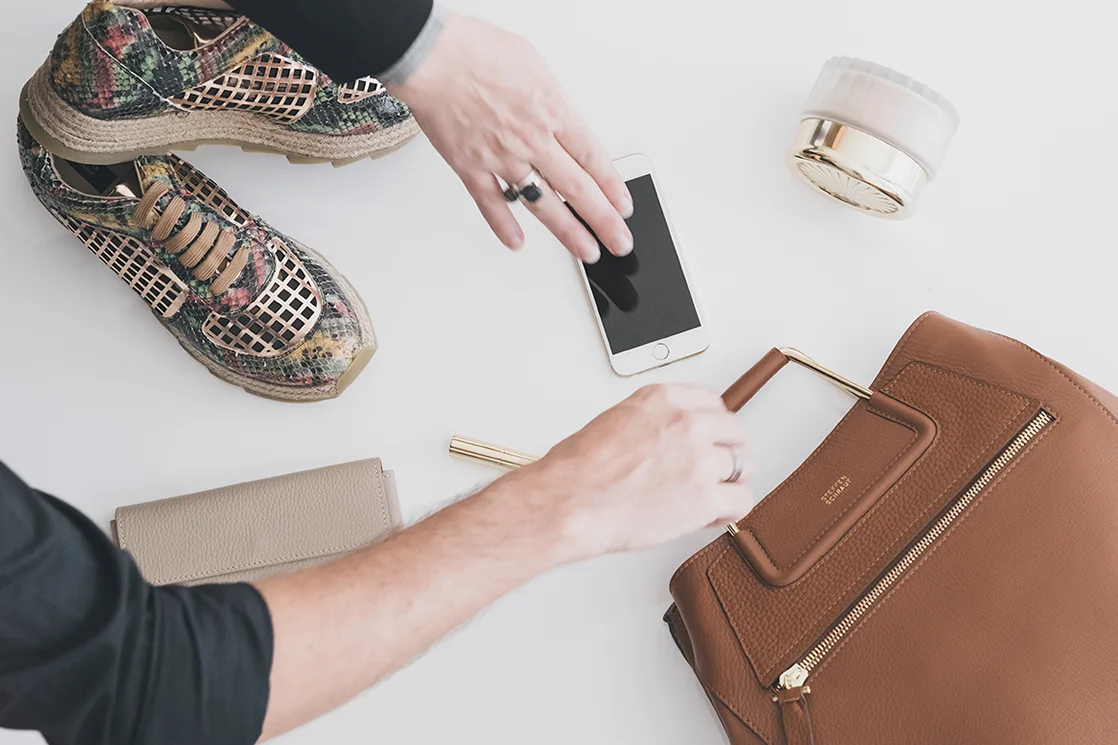 Verschiedene Gegenstände auf einem weißen Hintergrund: bunte Sneakers, eine braune Lederhandtasche, ein Smartphone, eine beige Geldbörse, ein Kosmetikprodukt in einer Dose und ein Stift. Hände greifen nach dem Smartphone.
