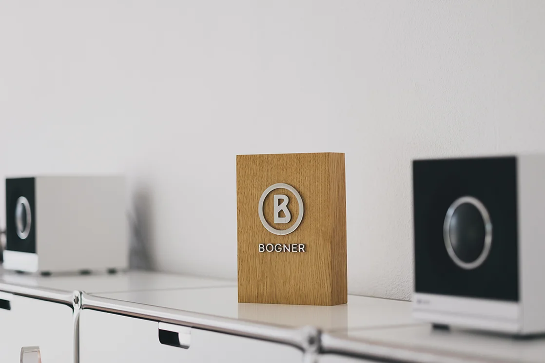Holzblock mit Logo und Schriftzug „BOGNER“, zwischen zwei Lautsprechern auf einem weißen Regal.
