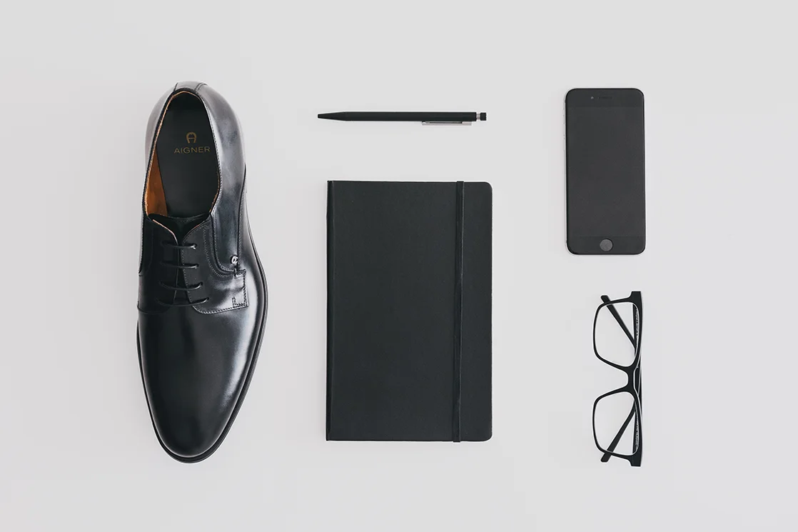 Flache schwarze Lederschuhe, ein schwarzes Notizbuch, ein schwarzer Stift, ein Smartphone und eine schwarze Brille auf weißem Hintergrund.
