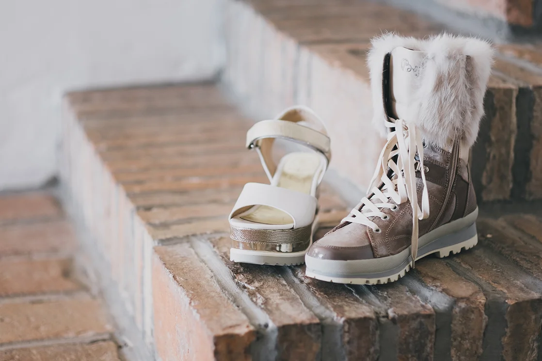 Ein Paar Schuhe auf einer Steintreppe - links ein eleganter High Heel-Sandalette in Beige mit Riemchen, rechts ein brauner Stiefel mit Fellbesatz und Schnürsenkeln.
