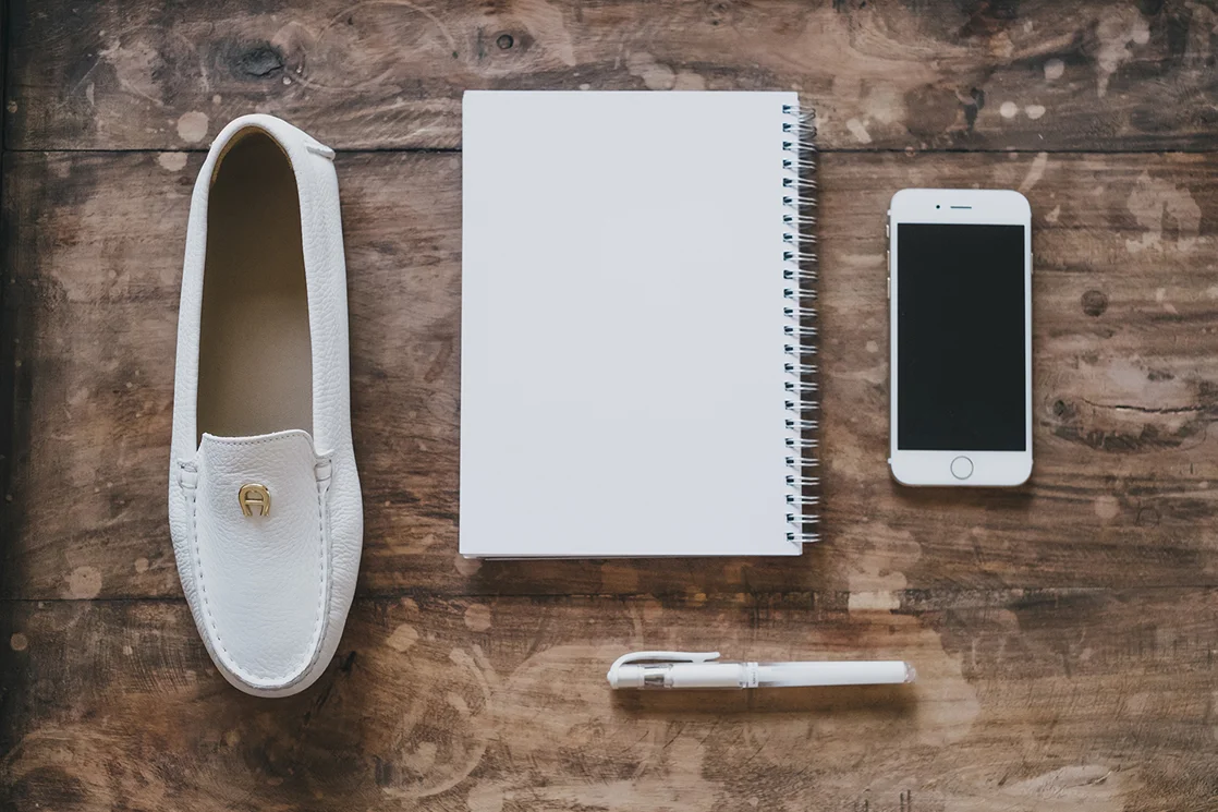 Weiße Loafer, Notizbuch, Stift und Smartphone auf einem Holztisch.