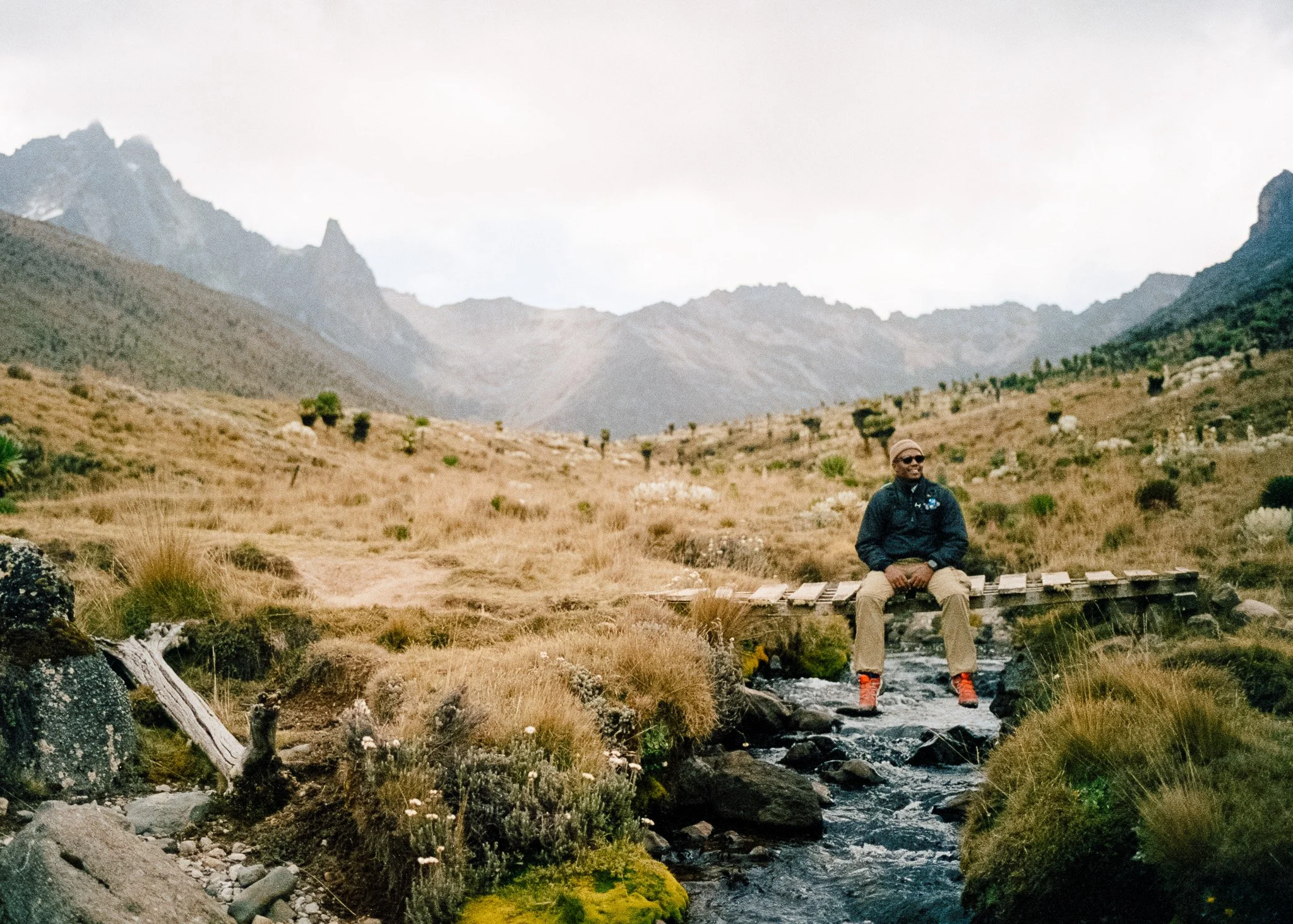 Person sitting on a small wooden bridge over a stream in a mountainous landscape with dry grass and scattered shrubs on MT. Kenya