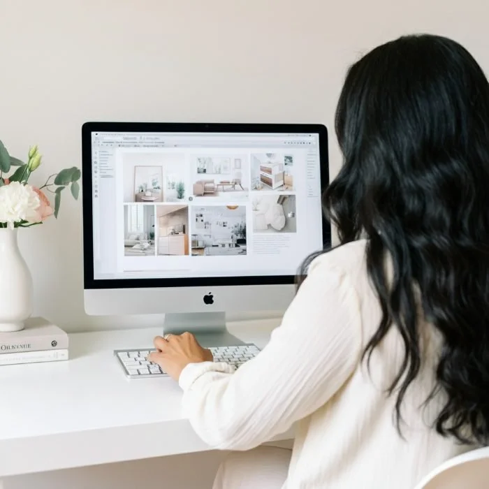 Person with long dark hair working on interior design photos on a computer in a bright, minimalist workspace with a white desk, computer, and a flower vase.