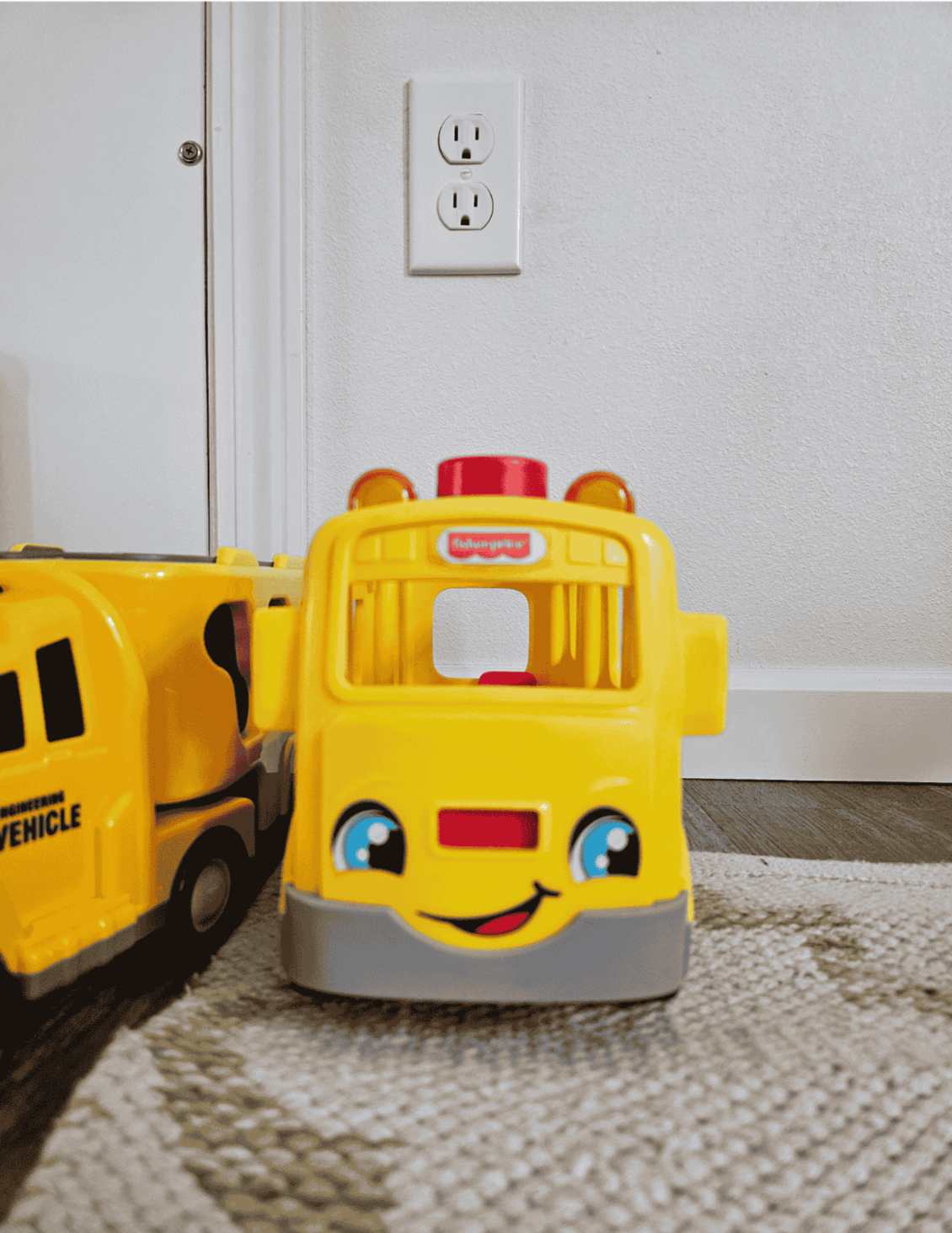 A child's toy, a yellow bus with a smiling face, sits on a carpet next to an electrical outlet.