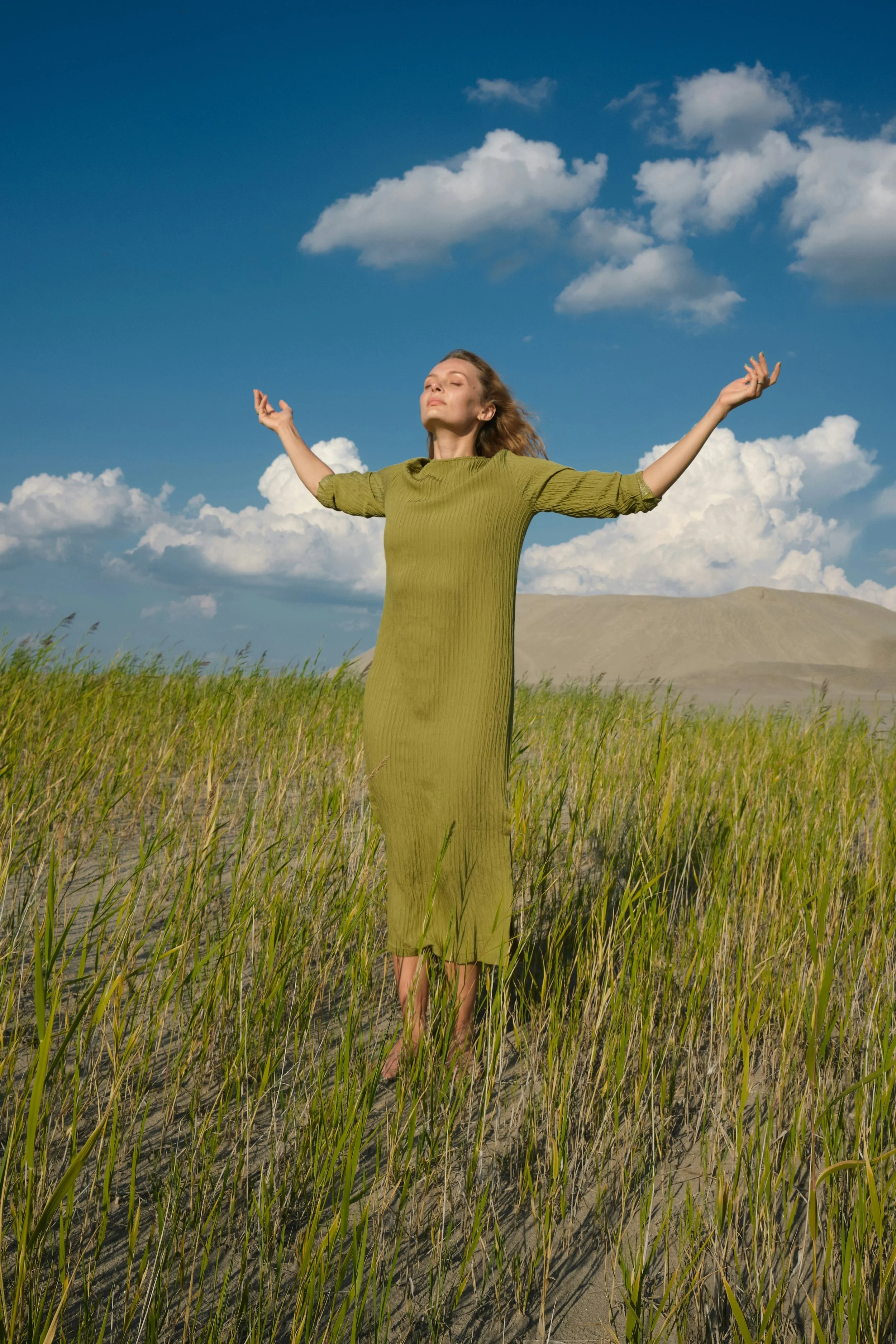 Mulher com vestido verde e cabelo ao vento, com os braços levantados, em um campo de grama sob céu azul com nuvens.