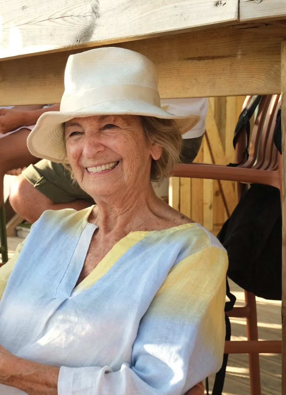 Mulher idosa sorrindo, usando chapéu de verão, unter de cores pastel, em ambiente com madeira, possivelmente ao ar livre.