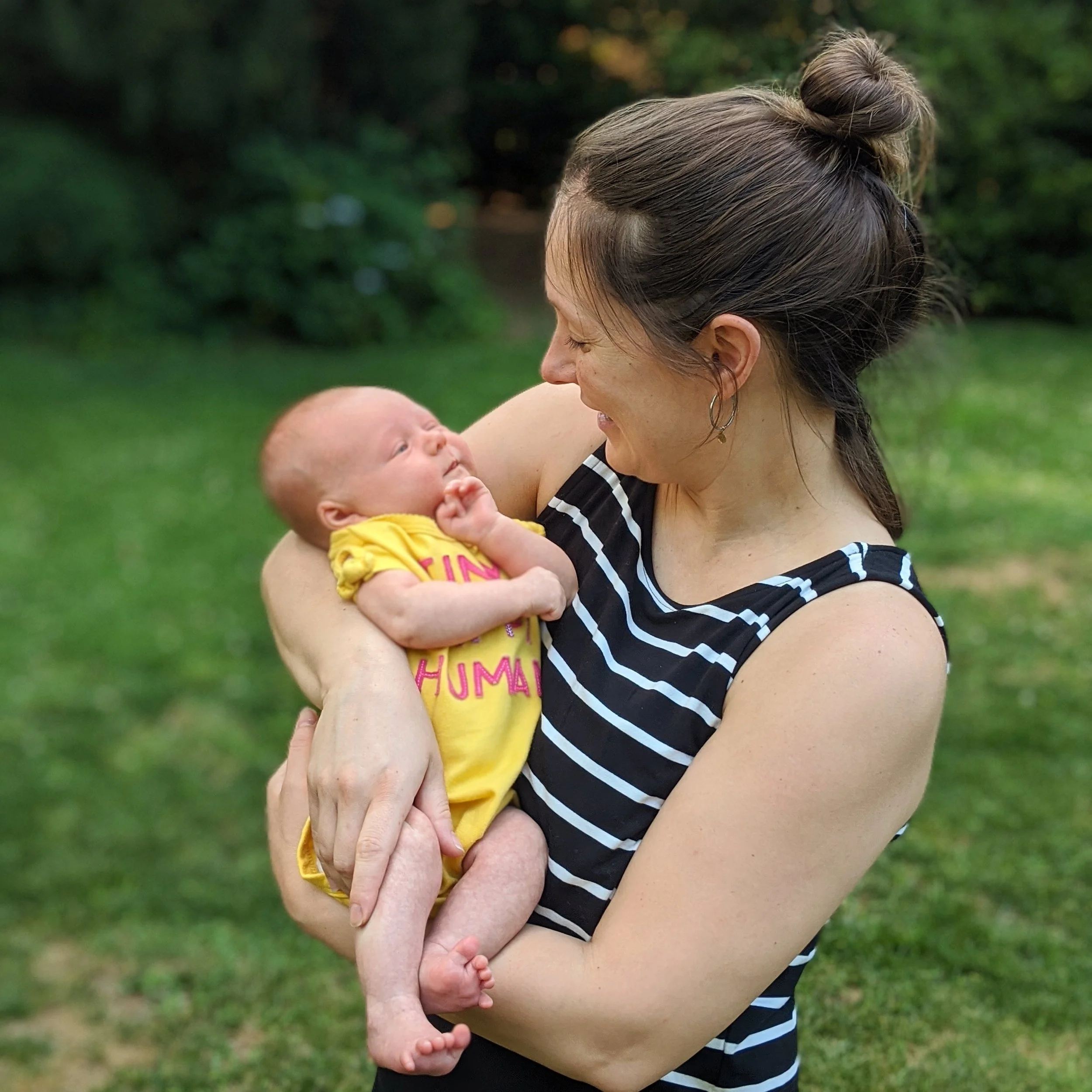 A woman holding a baby outdoors on a grassy area with green trees in the background. The woman is wearing a striped top and has her hair tied up in a bun. The baby is wearing a yellow outfit and looking up at the woman.