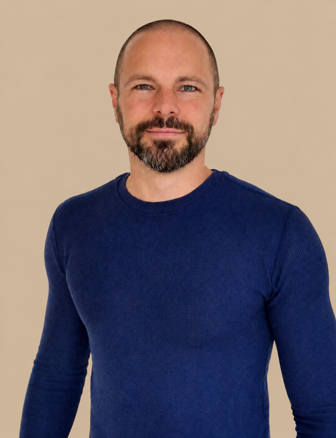 A man with a beard and short hair wearing a blue long-sleeve shirt standing against a plain beige wall.