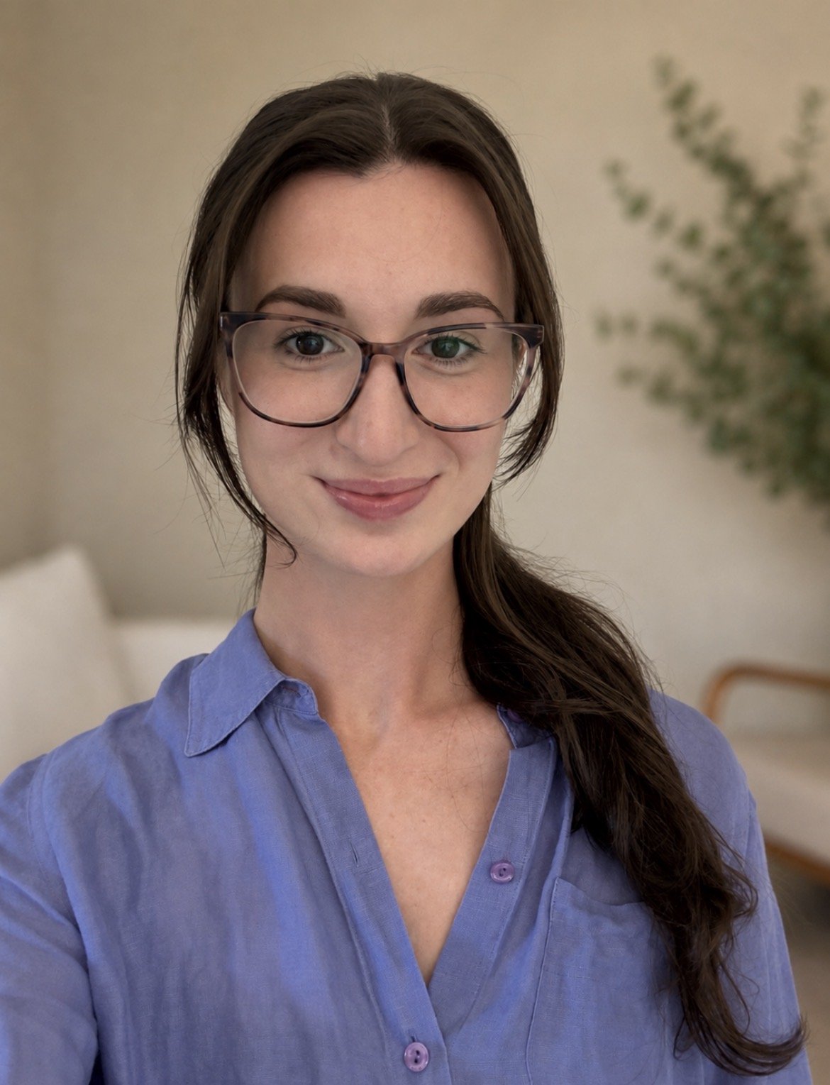 A woman with long brown hair, wearing glasses and a blue button-up shirt, smiling in a cozy, well-lit room.