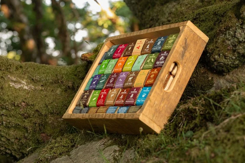 A wooden box containing an assortment of packaged soaps in colourful boxes, resting on a moss-covered tree branch outside.