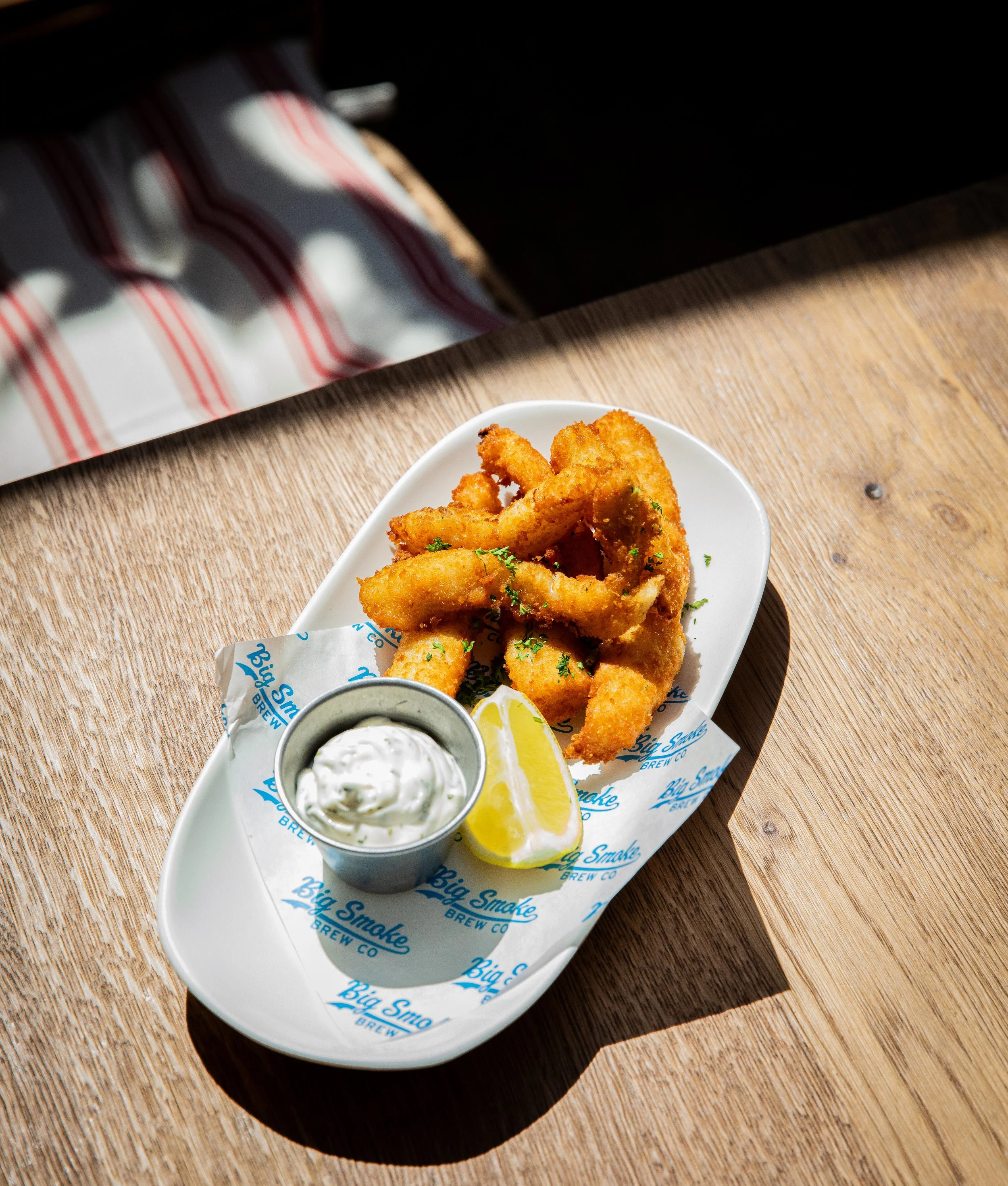 A plate of crispy fried calamari with a wedge of lemon and a side of tartar sauce, served on paper branded with 'Big Smoke Brew Co.' on a wooden table.