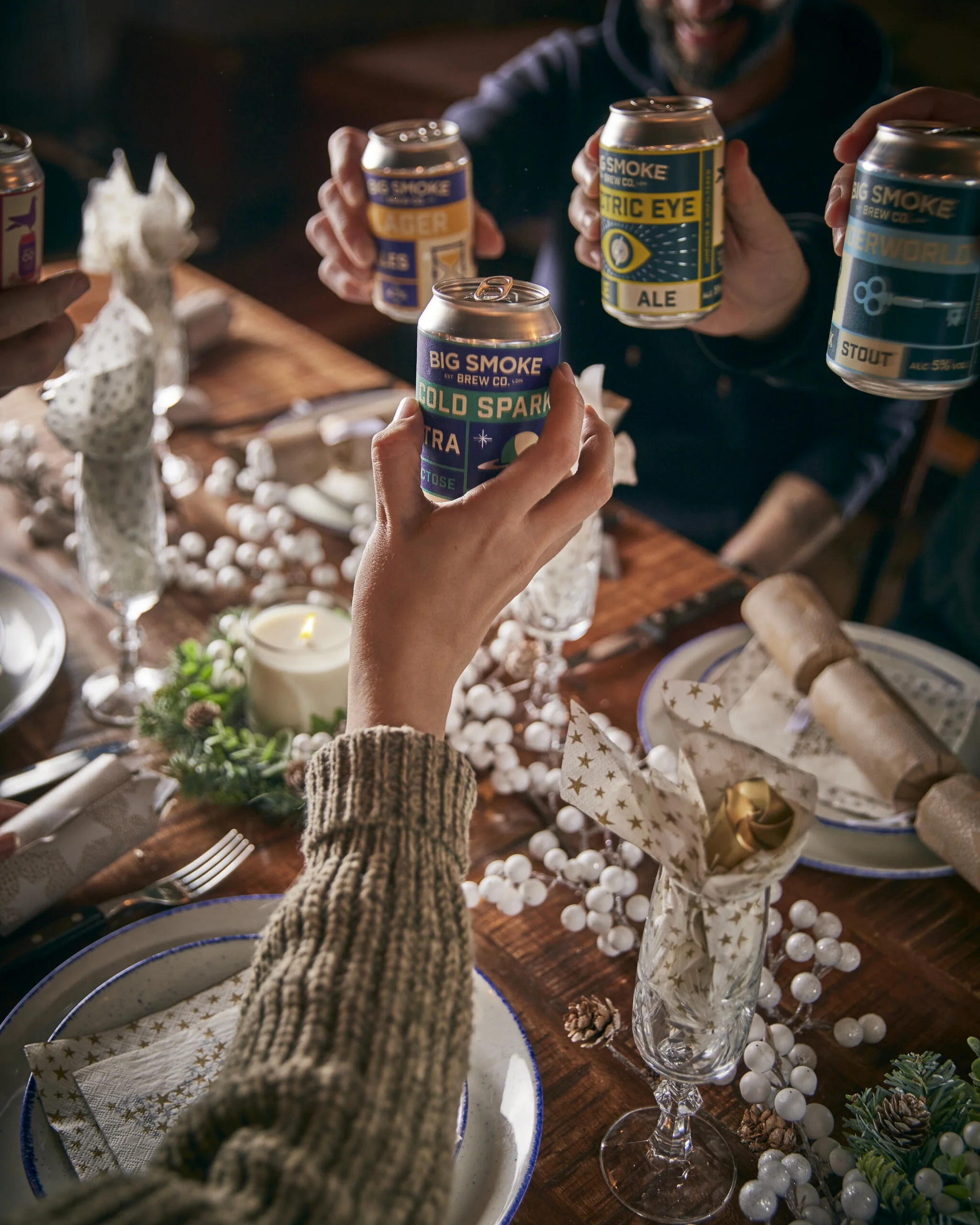 Group of people toasting with craft beer cans around a festive dinner table decorated with candles, greenery, and place settings.