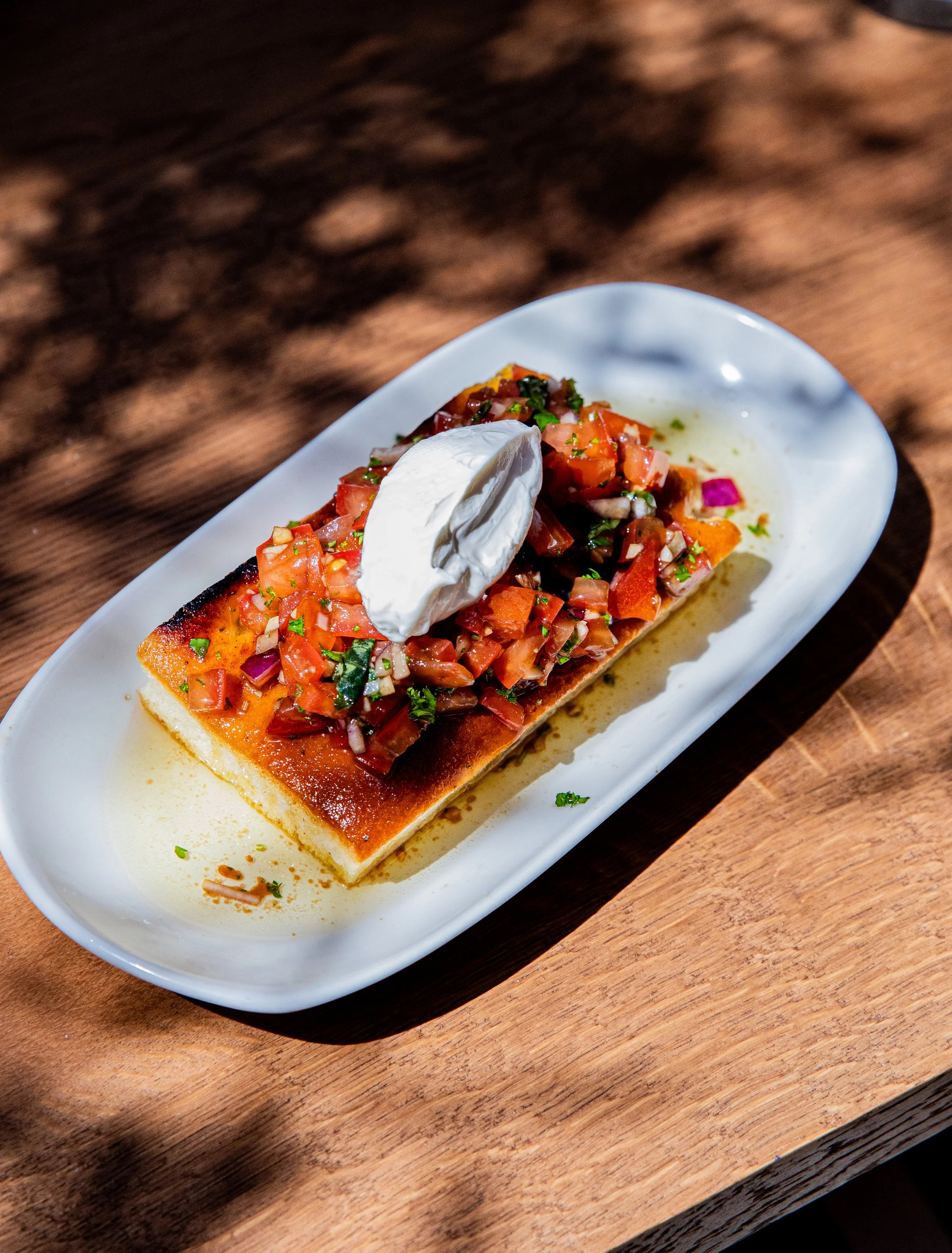 A piece of bread topped with diced tomatoes, onions, herbs, and a dollop of cream on a white plate.