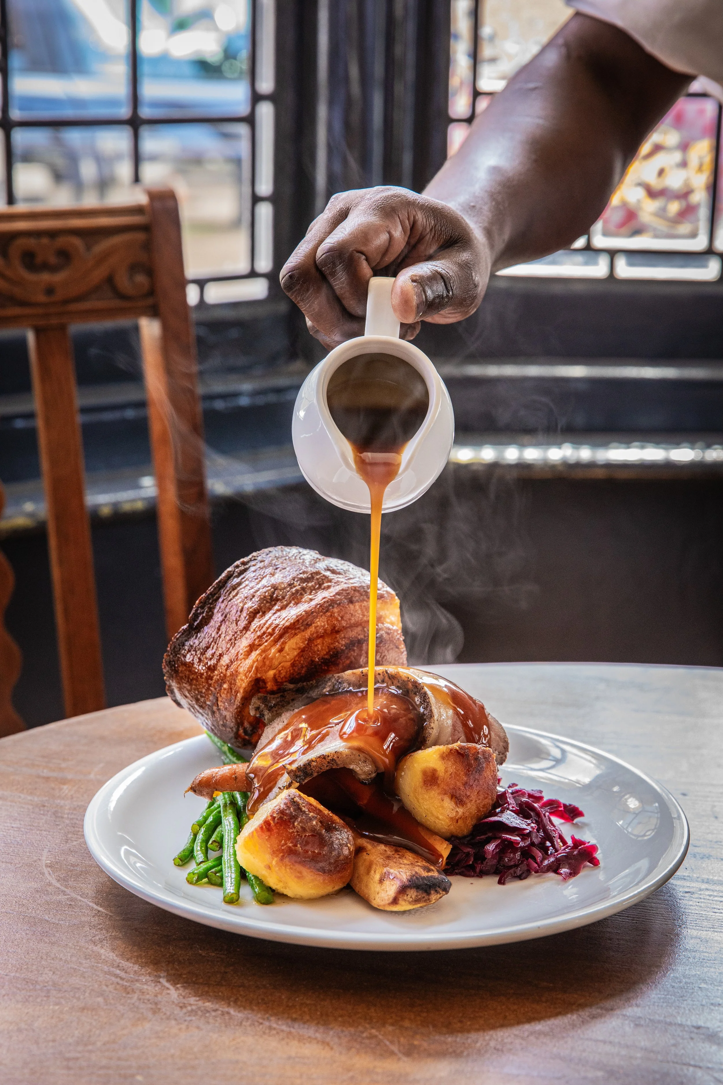 Sunday roast with beef, Yorkshire pudding, roasted potatoes, and carrots, being drizzled with gravy from a small pitcher.