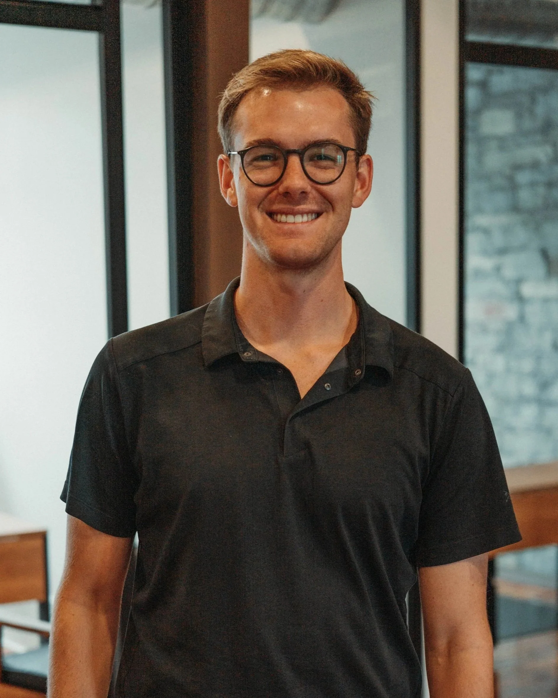 A young man with glasses, smiling, wearing a dark collared shirt, standing in front of a brick wall.