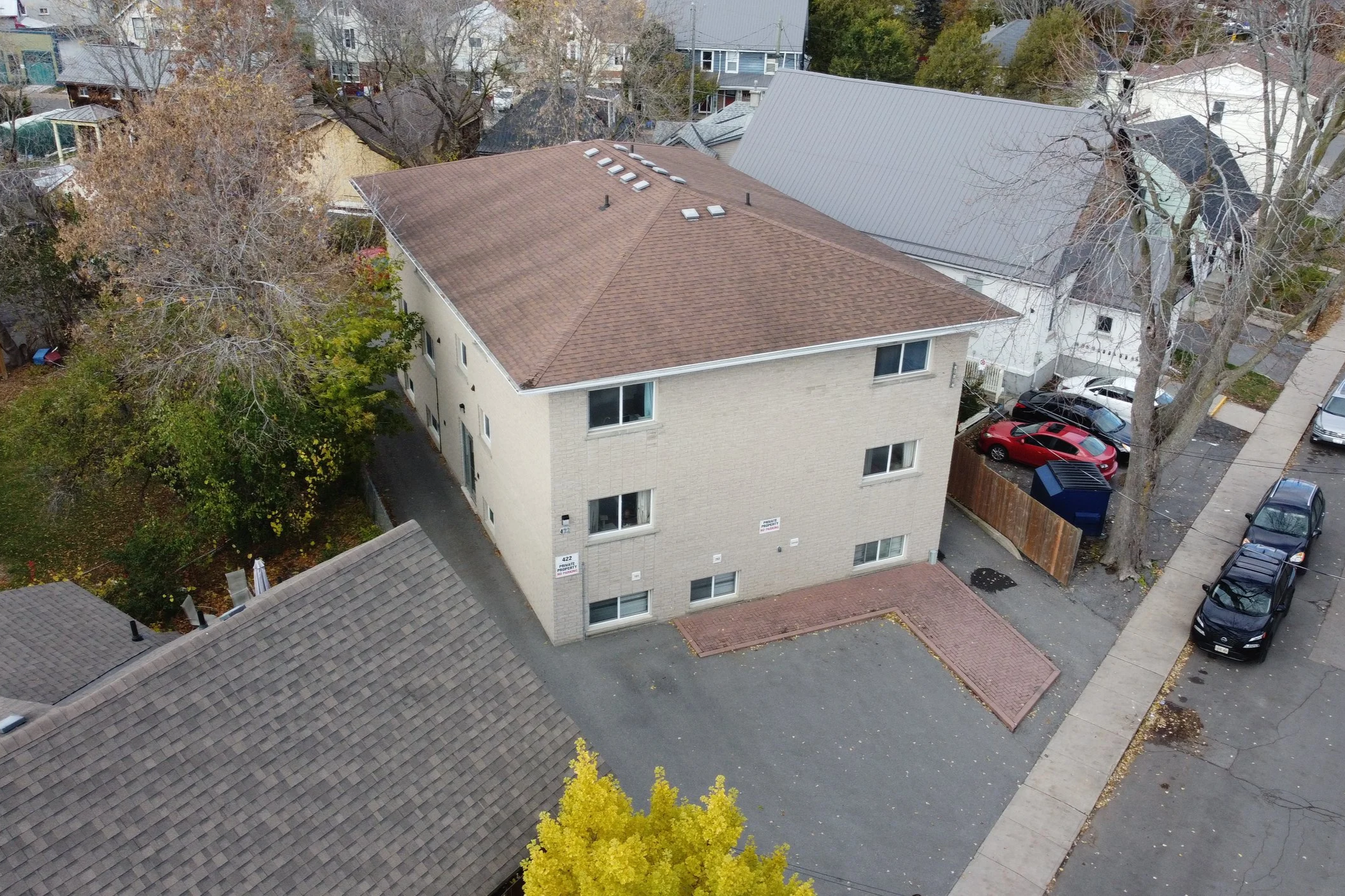 Aerial view of a three-story beige brick apartment building with a brown shingle roof, situated behind a parking lot and neighboring houses, surrounded by trees with some autumn foliage.