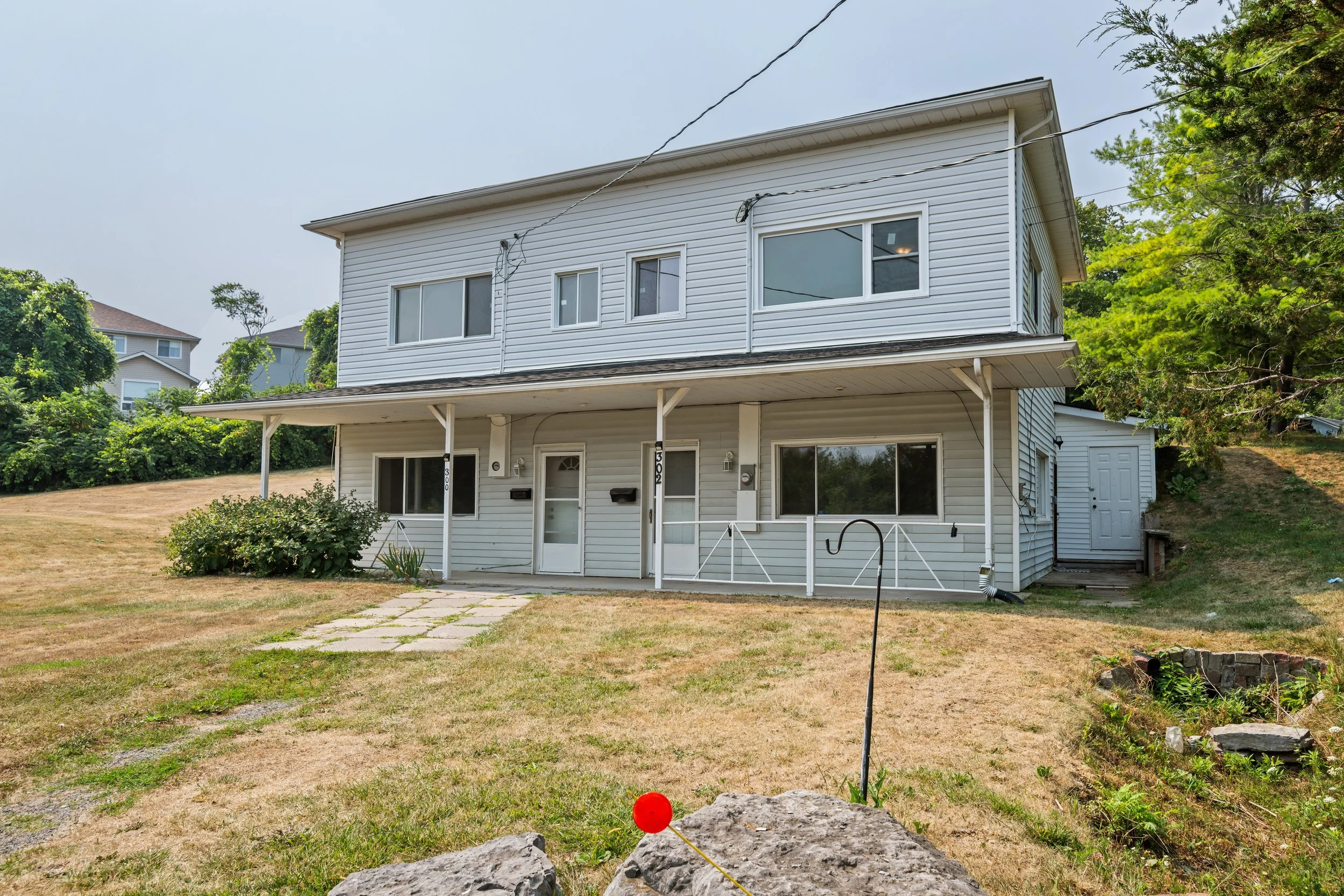 A two-story house with white siding, a front porch, and large windows, situated on a grassy lawn with a stone pathway leading to the front door, surrounded by trees and neighboring houses.