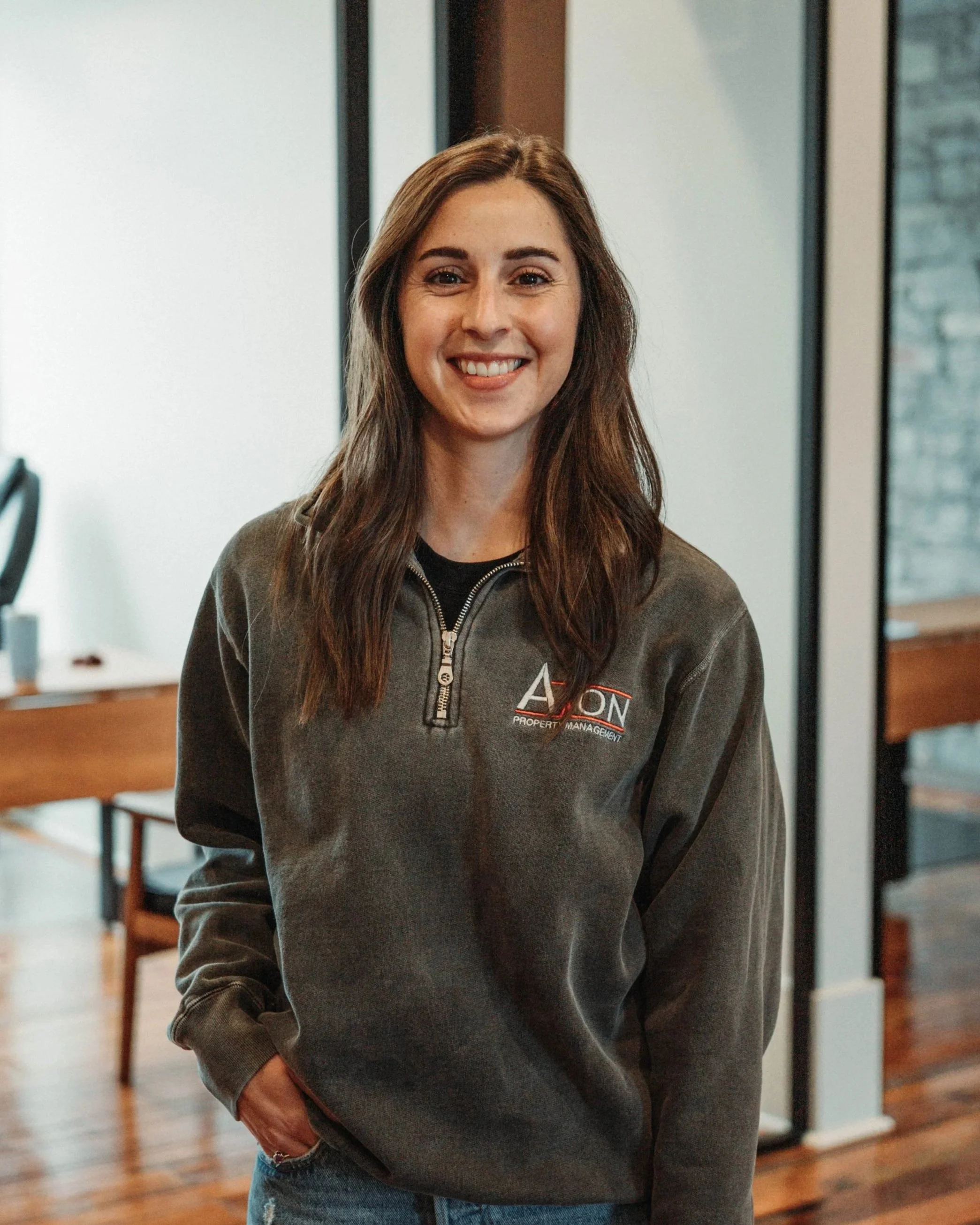 A young woman with long wavy hair smiling, wearing a dark top and a light jacket, standing in front of a textured stone wall.